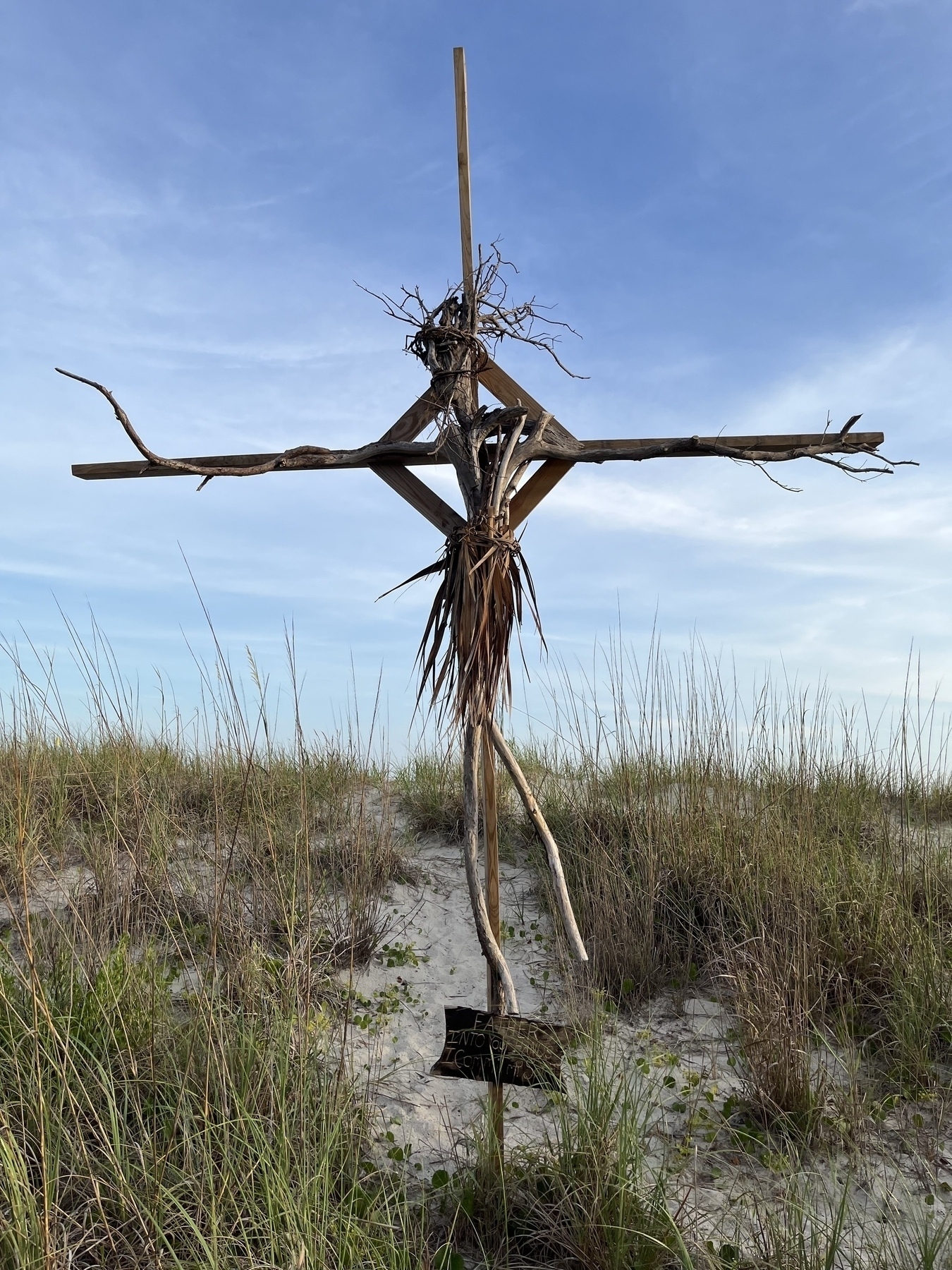 A wooden stick figure resembling a Jesus on the cross stands in a grassy sandy area under a blue sky.