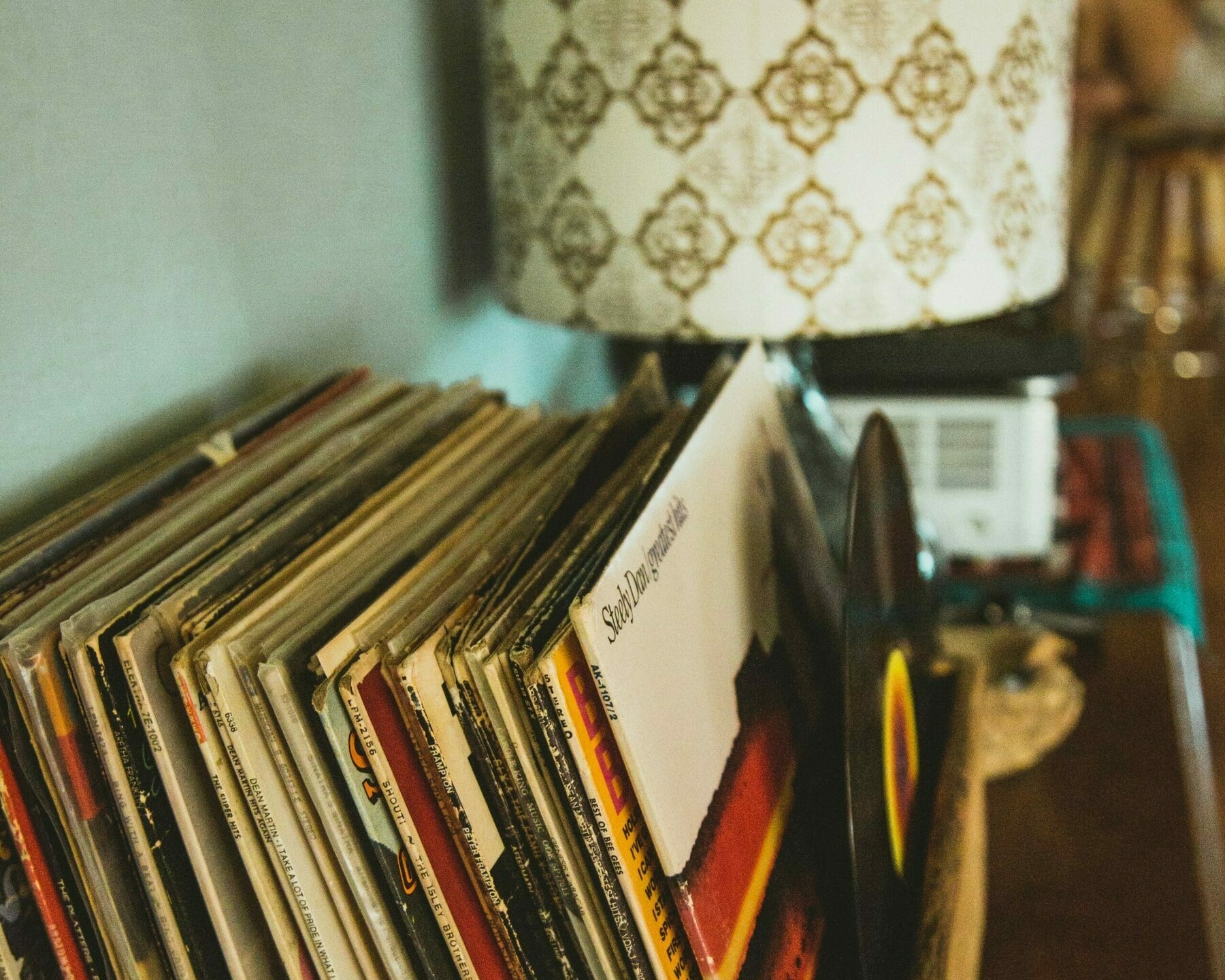 Auto-generated description: A collection of vinyl records is stacked on a shelf next to a patterned lamp and a record player.