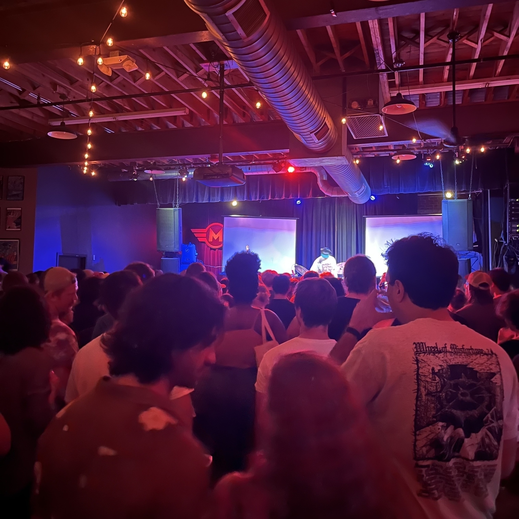 A large crowd of people is gathered indoors, facing a stage with a DJ, under a ceiling decorated with exposed beams and string lights.