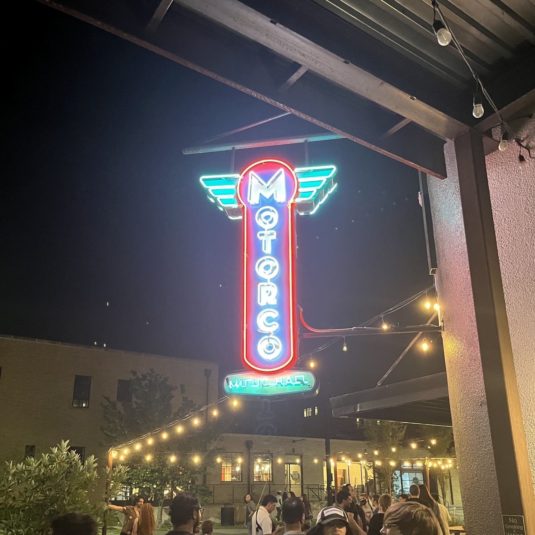 A neon sign reading "Motorco Music Hall" is illuminated, hanging above a crowd and surrounded by string lights at night.