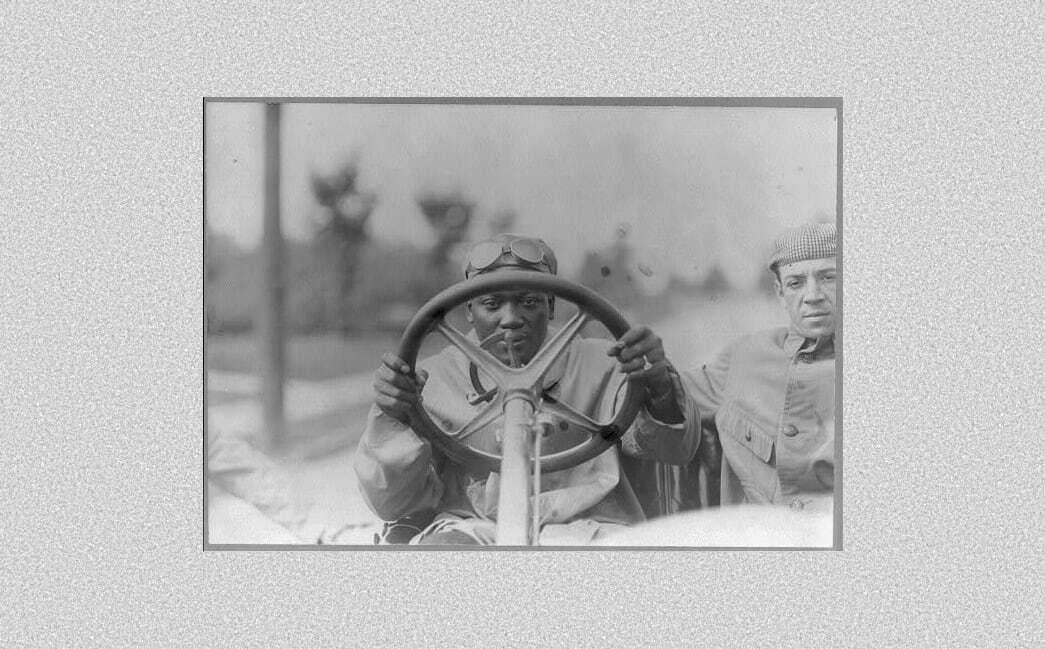 Auto-generated description: Two people are sitting in a vintage car, with one of them holding the steering wheel.